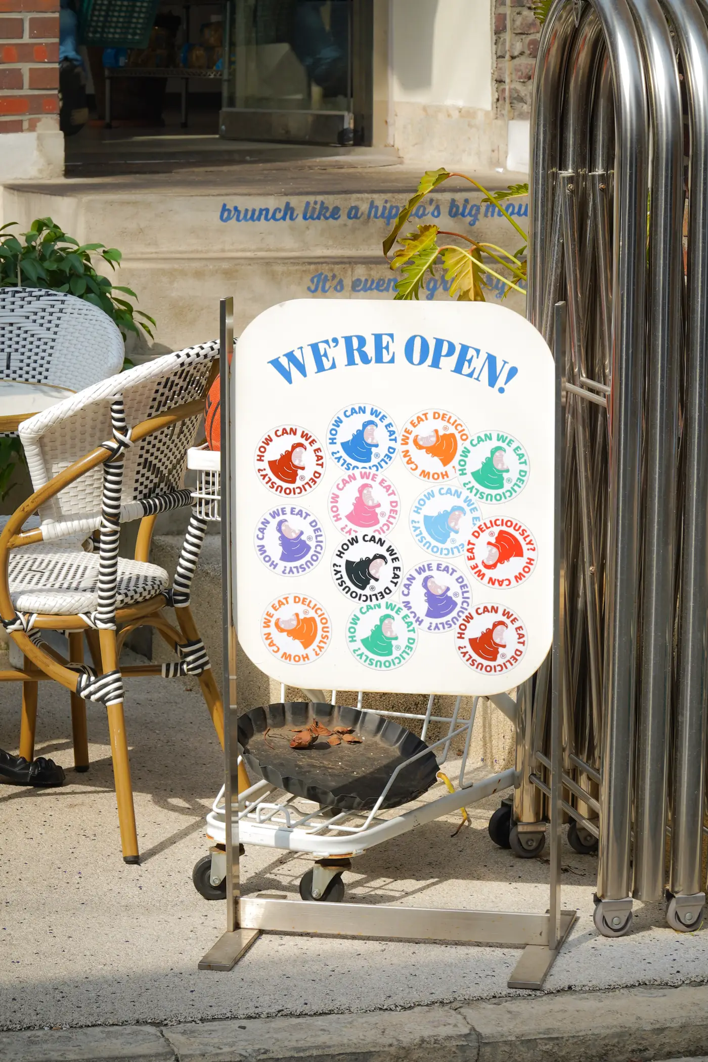 'WE'RE OPEN!' 입간판과 라탄 의자가 놓인 히포 브런치하우스 테라스, 벽에 적힌 'brunch like a hippo's big mouth'